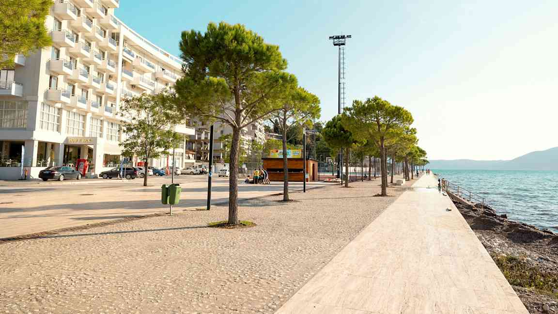 En strandpromenade med trær og hvitflislagt gulv langs vannet, med en moderne bygning til venstre. (AI-generert tekst)