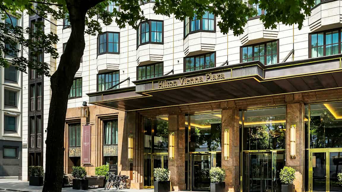 En hotellbygning med en elegant inngang, omgitt av trær. (AI-generert tekst)
