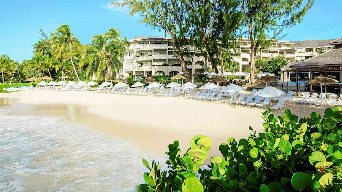 En strand med solsenger og parasoller foran en resort med palmer og beplantning. (AI-generert tekst)