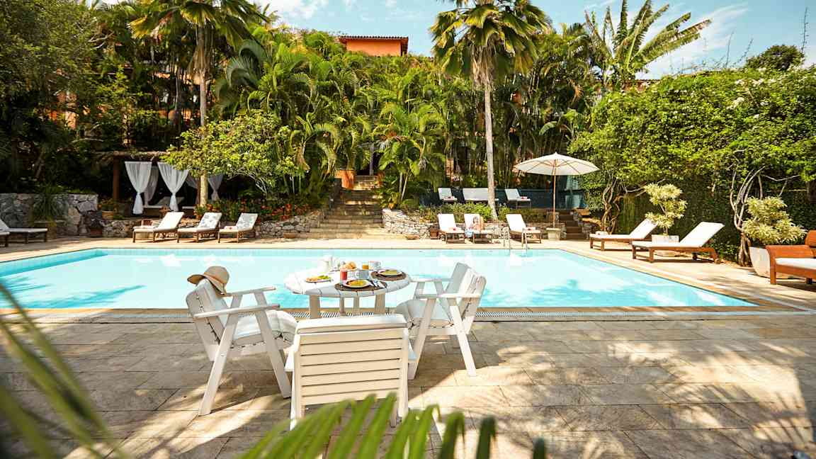 Et utendørs basseng omgitt av frodig vegetasjon og solstoler. Et bord med mat og drikke står ved bassengkanten. (AI-generert tekst)