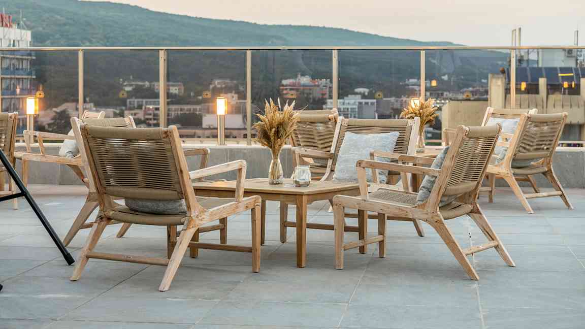 Terrasse med en gruppe tremøbler, inkludert stoler og bord. Utsikt over byen og grønne åser i bakgrunnen. (AI-generert tekst)