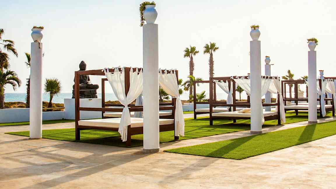 Utendørsområde med baldakinsenger og statue ved sjøen. (AI-generert tekst)