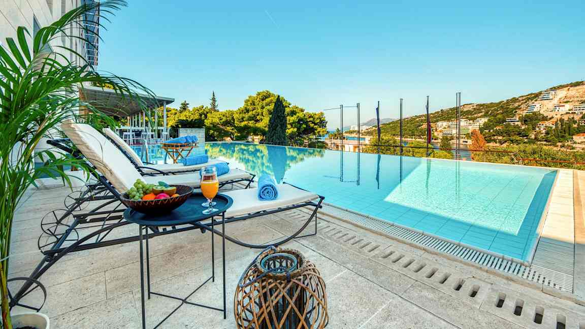 En utsikt mot et infinitybasseng med solsenger, bord med frukt og drikke ved siden av, samt grønne omgivelser og himmel i bakgrunnen. (AI-generert tekst)