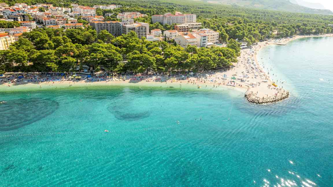 Luftbilde av en kyst med klart blått vann, strand med solsenger og tett bebyggelse omgitt av skog og fjell. (AI-generert tekst)