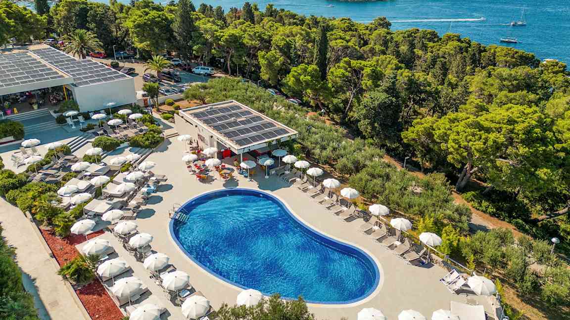 Utsikt over et utendørsbasseng omgitt av solsenger med parasoller, plassert nær kysten med utsikt over skog og hav. (AI-generert tekst)