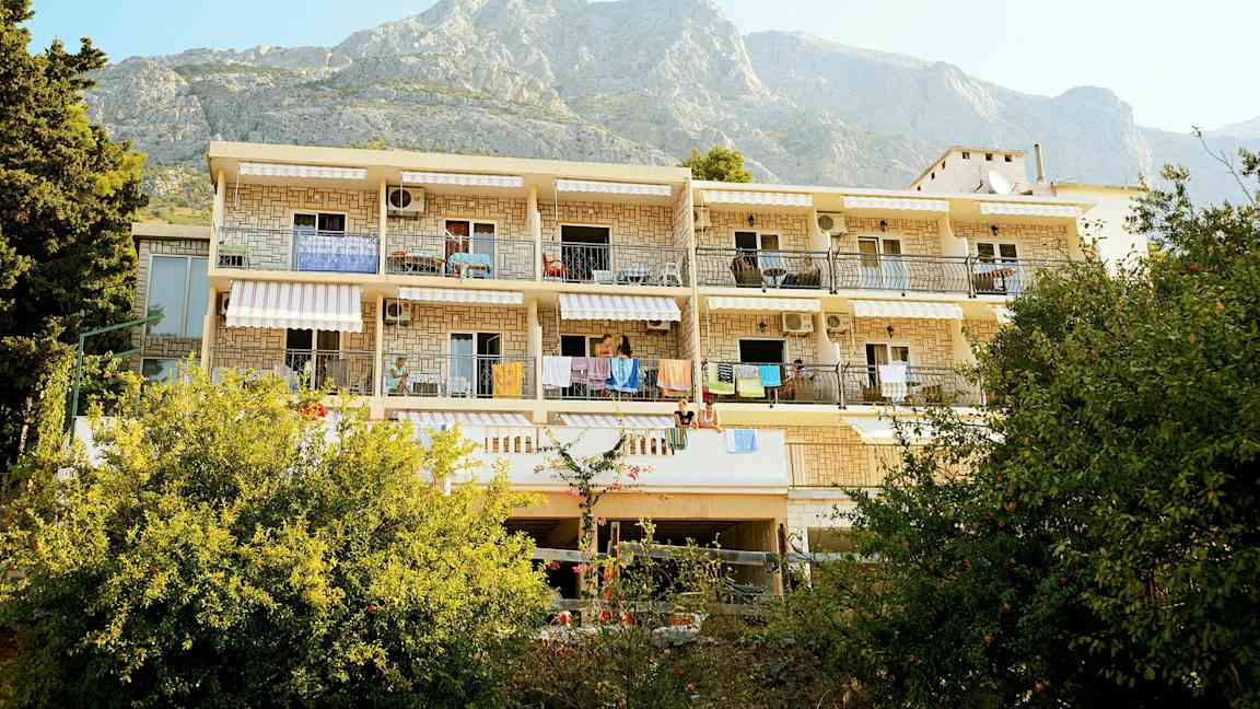 En bygning med balkonger og hengende håndklær, omgitt av grøntområder og med fjell i bakgrunnen. (AI-generert tekst)