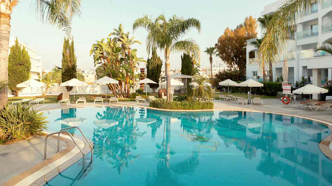 Utendørs svømmebasseng omgitt av solsenger og parasoller, med palmer og bygninger i bakgrunnen. (AI-generert tekst)