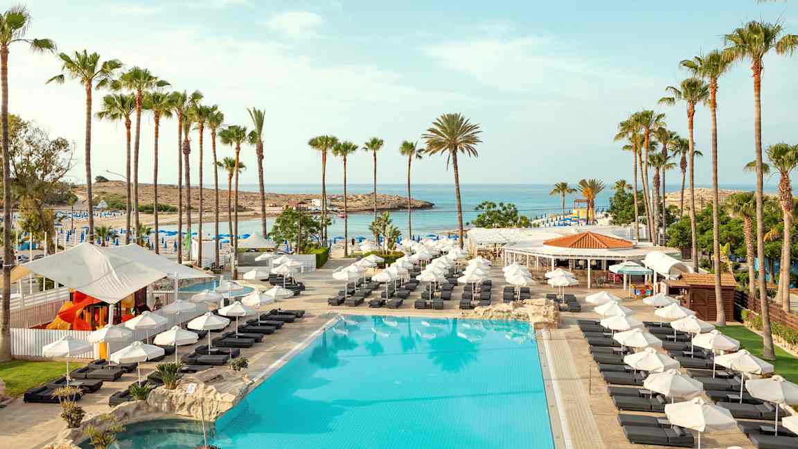 Bilde av et basseng med solsenger, parasoller og palmer ved en strand. (AI-generert tekst)