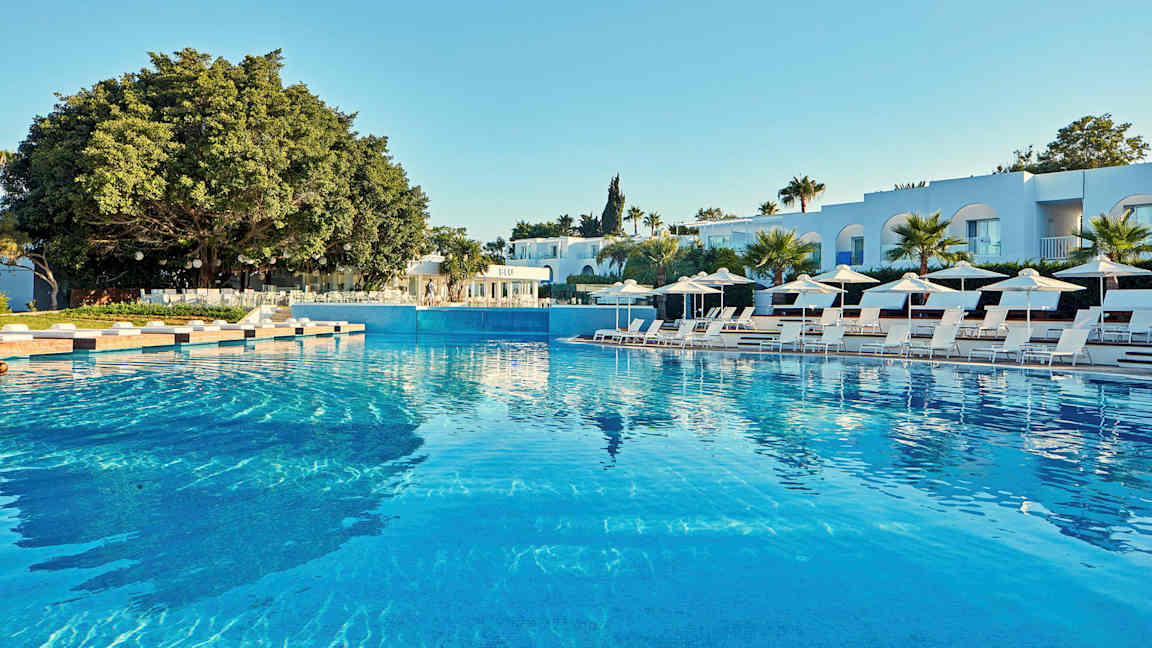 Bilde av et stort basseng omgitt av solsenger, parasoller og moderne bygninger i bakgrunnen. (AI-generert tekst)