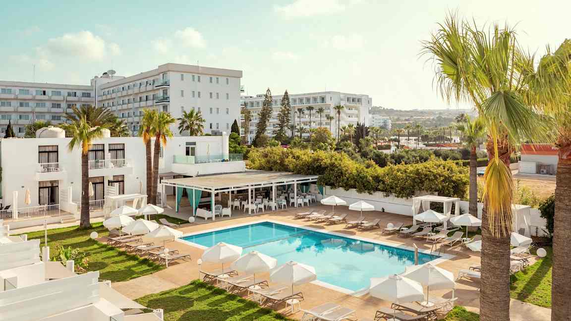 Et hotellområde med basseng, solsenger og parasoller, omgitt av palmer og bygninger. (AI-generert tekst)