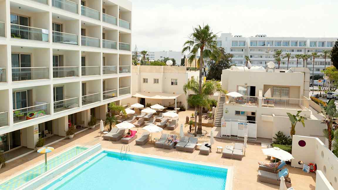 Hotellområde med basseng, solsenger og bygninger. (AI-generert tekst)