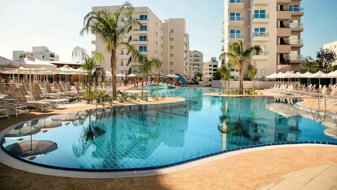 Et utendørs svømmebasseng omgitt av solsenger og parasoller, med høye bygninger i bakgrunnen. (AI-generert tekst)