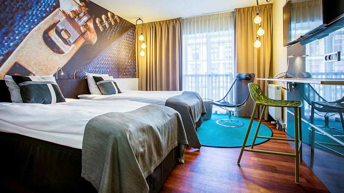 Et moderne designet hotellrom med to enkeltsenger, en illustrert veggdekorasjon, gardiner og vindusutsikt. Rommet har også en grønn stol, en svart stol og en veggmontert TV. (AI-generert tekst)