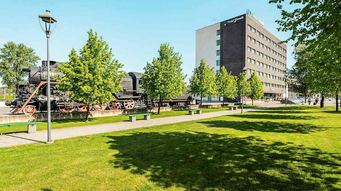 Et gammelt damplokomotiv står utendørs ved siden av et moderne hotellanlegg og frodige trær. (AI-generert tekst)