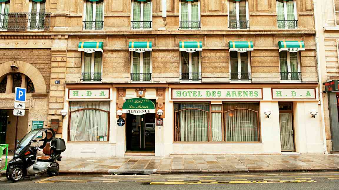 Bygning med hotellfasade, skilt med teksten 'Hotel des Arenes' og grønnhvite markiser over vinduene. (AI-generert tekst)