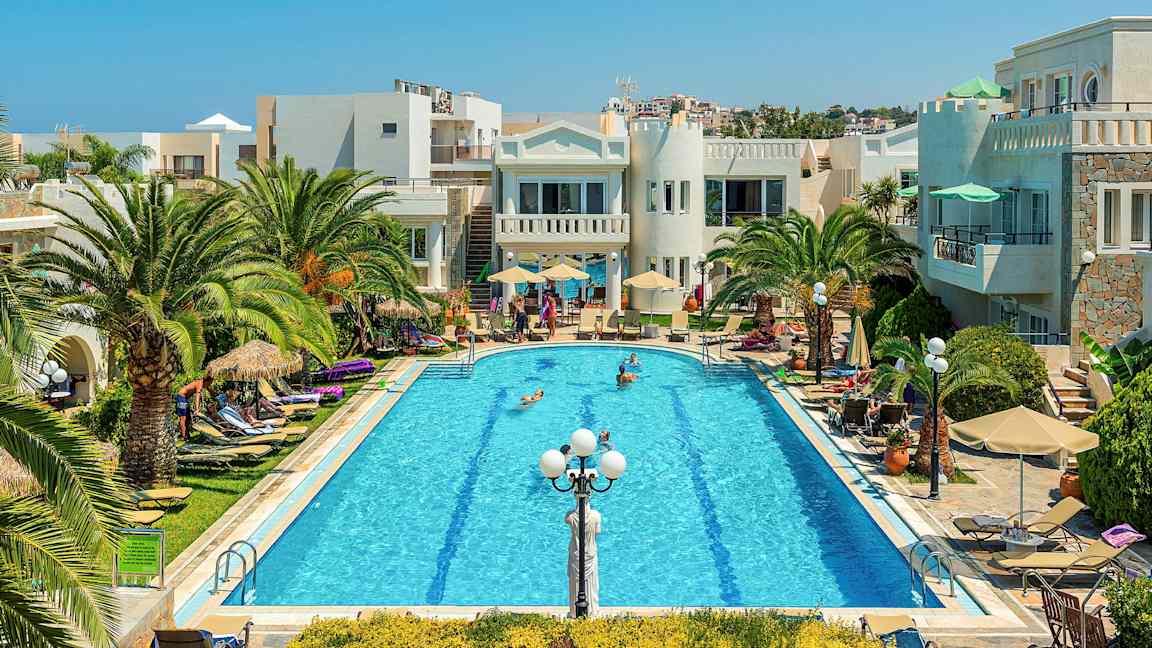 Et basseng omgitt av palmer, solsenger og bygninger under en klar blå himmel. (AI-generert tekst)