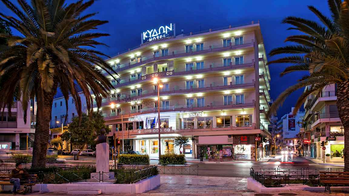 En bygning med flere etasjer, muligens et hotell, i en by eller bymiljø om kvelden, omgitt av palmer og gatelys. (AI-generert tekst)