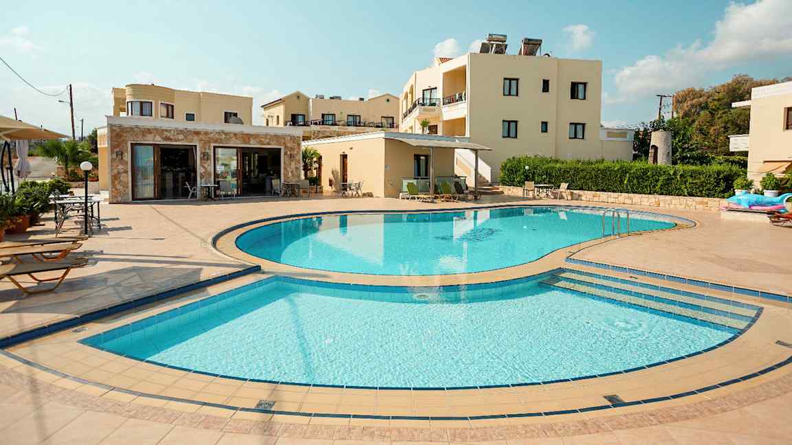 Et bilde som viser et utendørsbasseng omgitt av solsenger og bygninger i bakgrunnen. (AI-generert tekst)