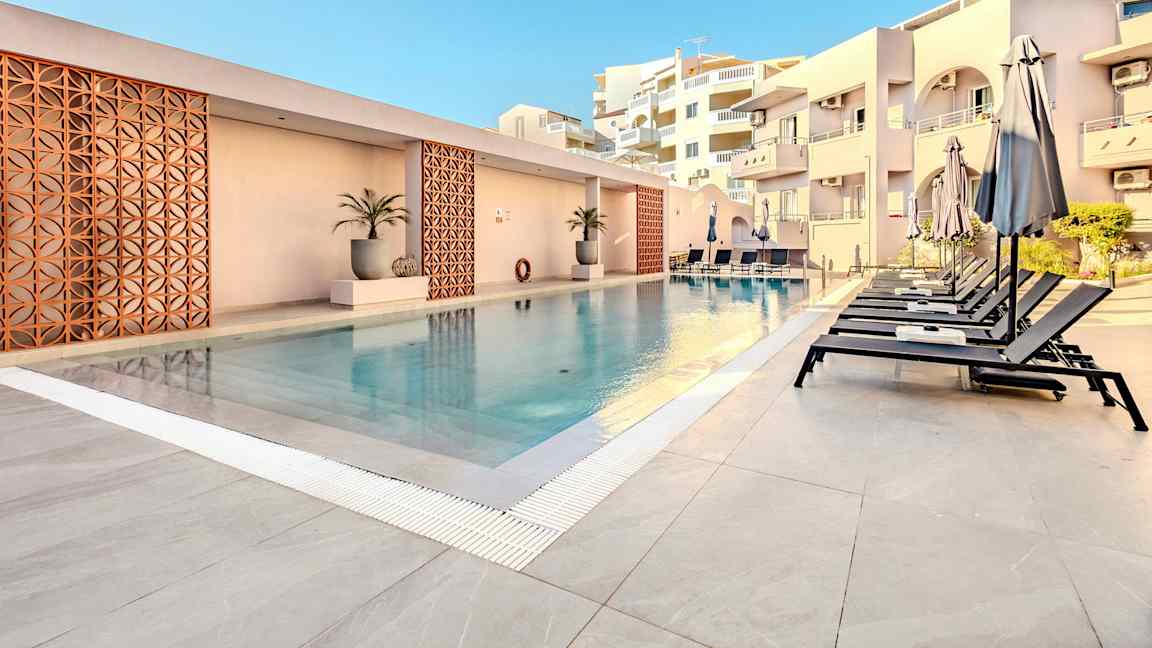 Et utendørs svømmebasseng med solsenger og parasoller ved siden, omgitt av bygninger. (AI-generert tekst)