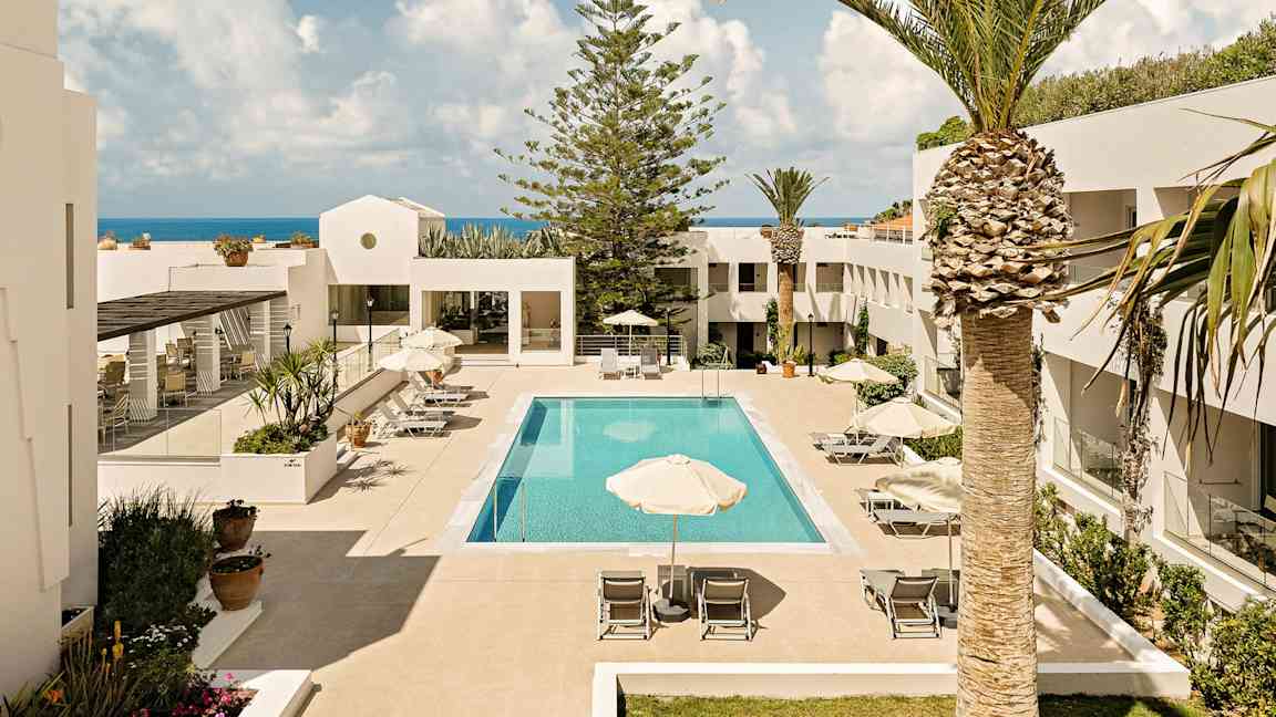 Hotellets atrium med et basseng omgitt av solsenger og parasoller, innrammet av hvite bygninger og palmer. Havet kan ses i bakgrunnen. (AI-generert tekst)