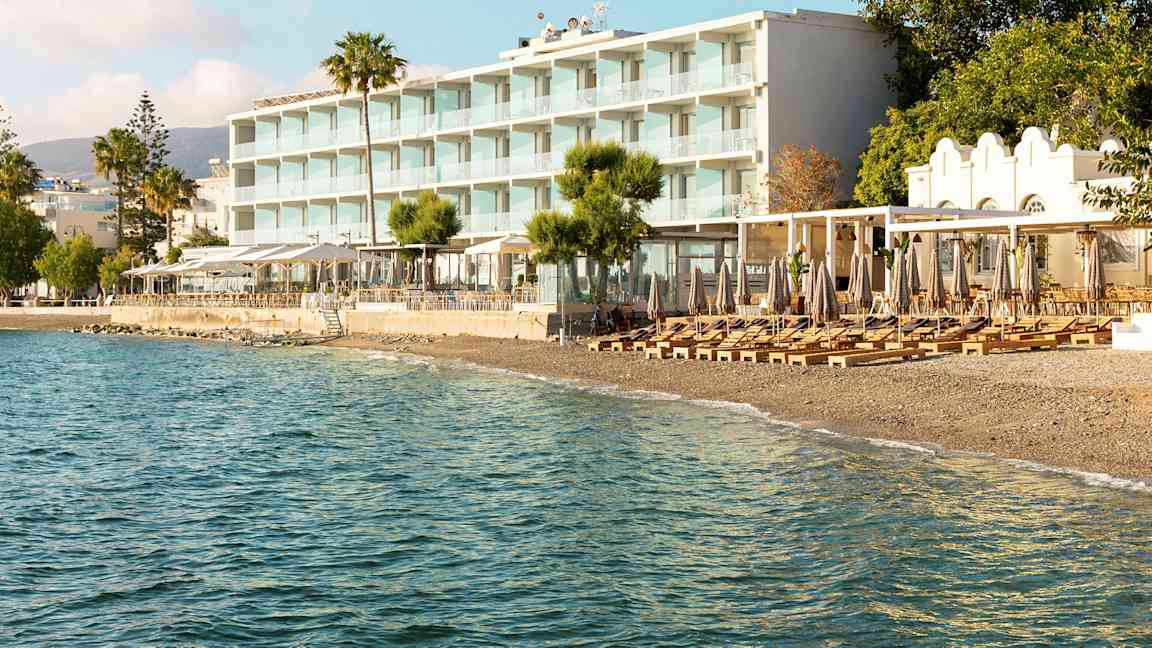 Utsikt mot et strandhotell med solsenger og parasoller på stranden og havet i forgrunnen. (AI-generert tekst)