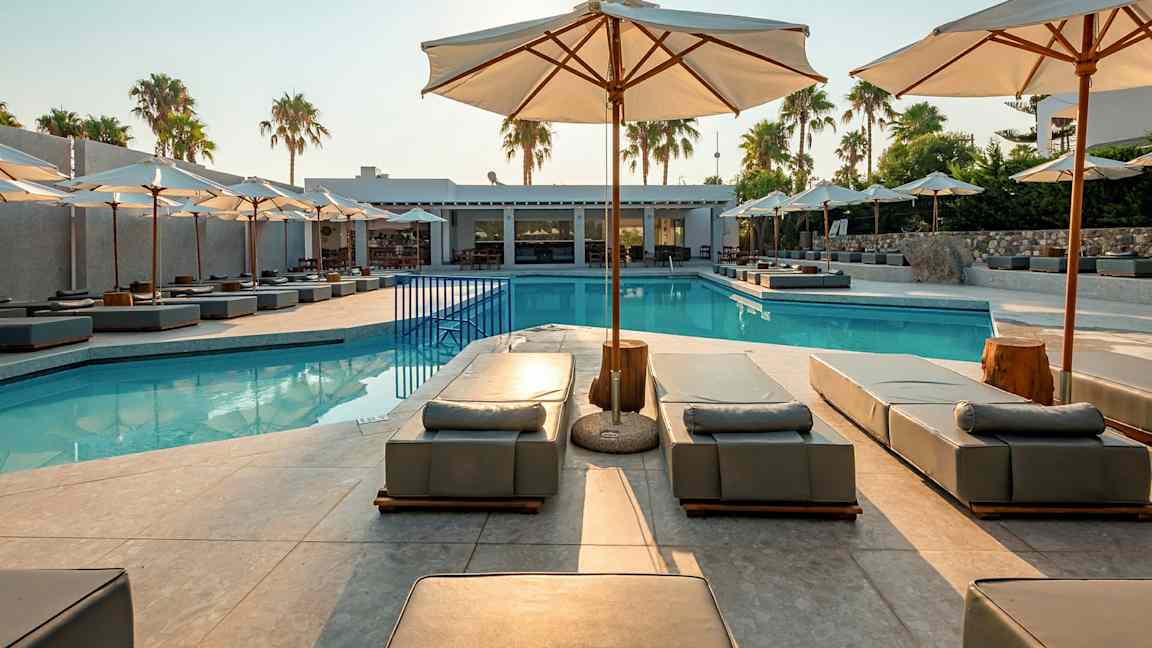Utendørsbasseng med solsenger og parasoller, omgitt av palmer og bygninger. (AI-generert tekst)