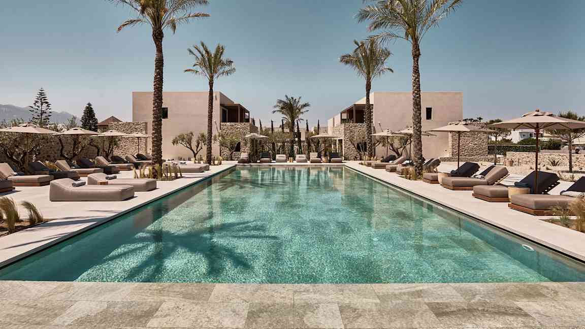 Et rektangulært utendørs svømmebasseng omgitt av solsenger og parasoller, med palmer og bygninger i bakgrunnen. (AI-generert tekst)