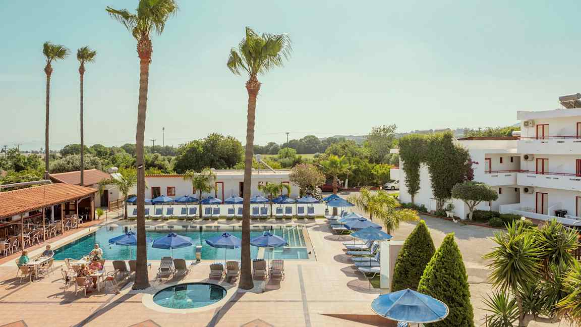 Et basseng med solsenger og parasoller omgitt av bygninger og trær. (AI-generert tekst)