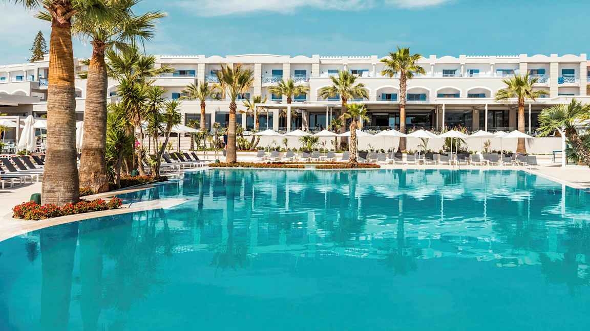 Et stort basseng foran et hotell med palmer rundt og en blå himmel over. (AI-generert tekst)