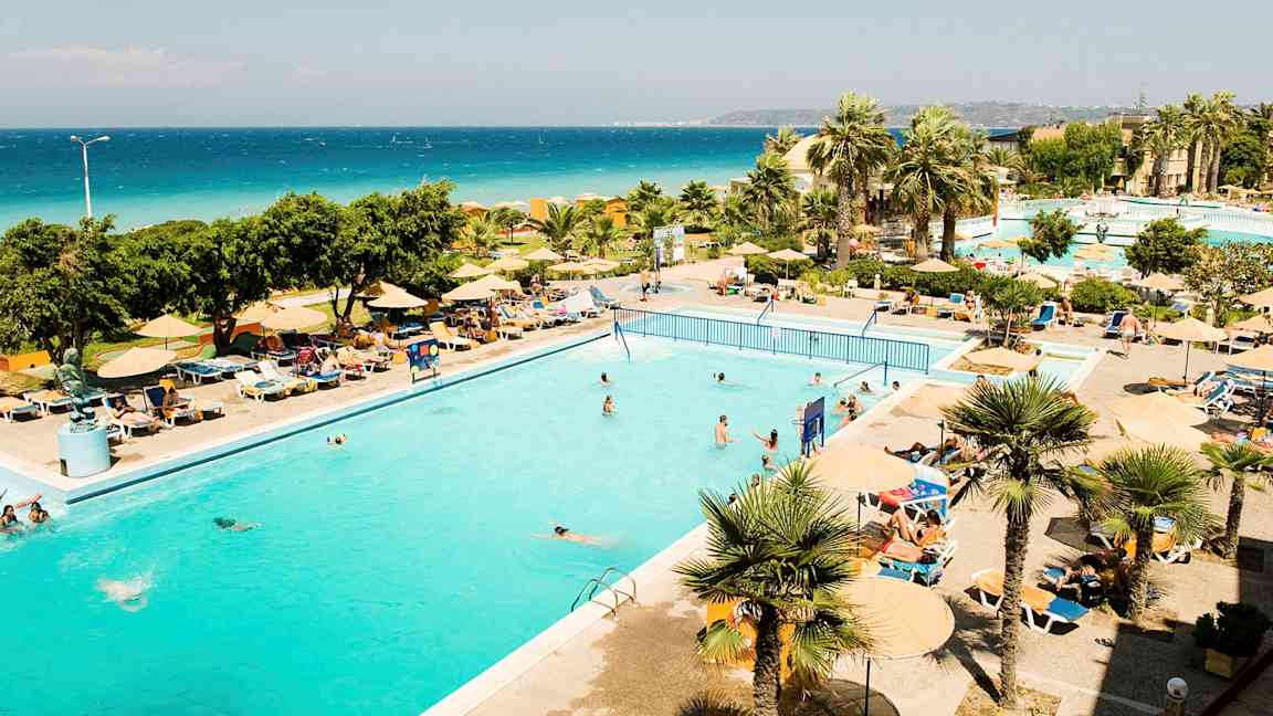 Et utendørsbasseng omgitt av solsenger og parasoller, med palmer og havet i bakgrunnen. (AI-generert tekst)