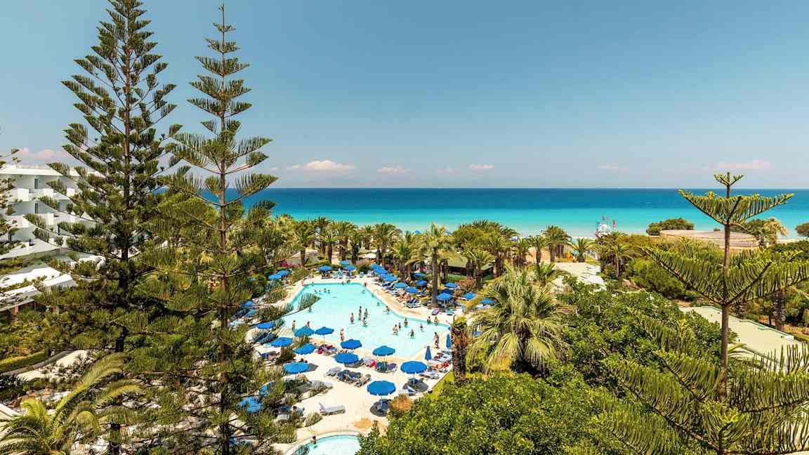 Bilde av hotellområde med basseng og solsenger omgitt av grøntområder og utsikt mot havet. (AI-generert tekst)