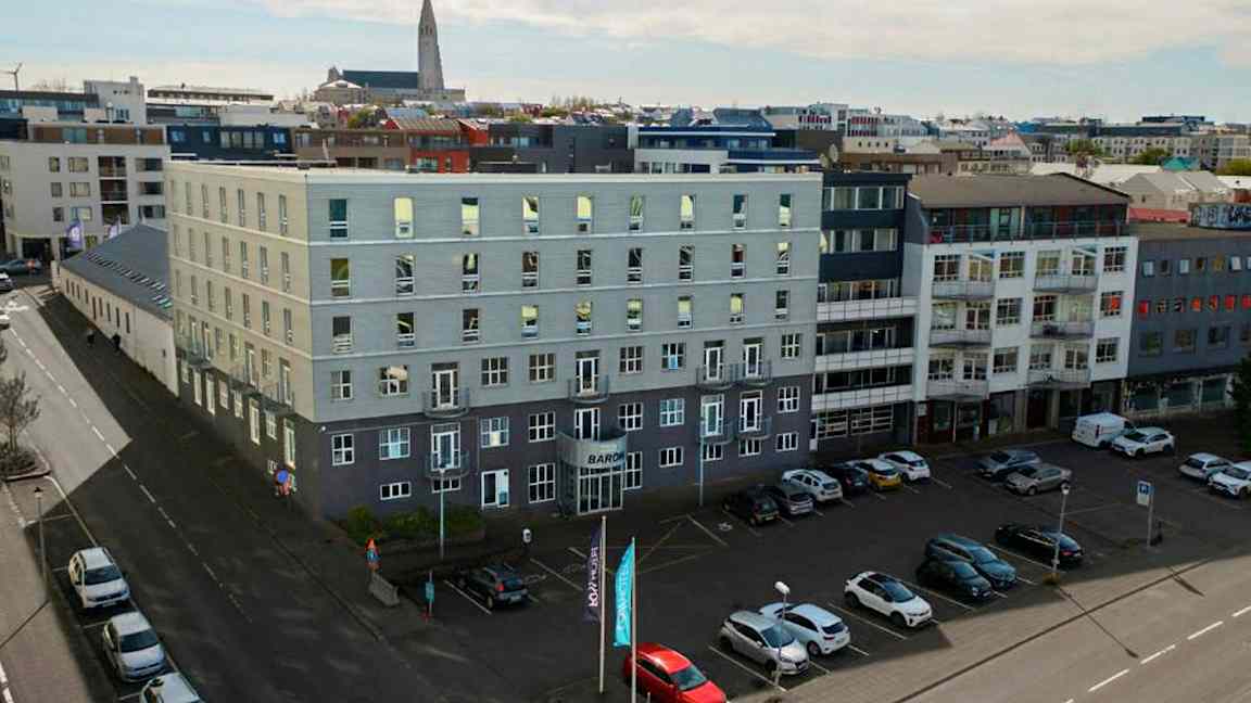 Flere moderne bygninger omgir et åpent parkeringsområde, med biler parkert på bakken og en kirke synlig i bakgrunnen. (AI-generert tekst)