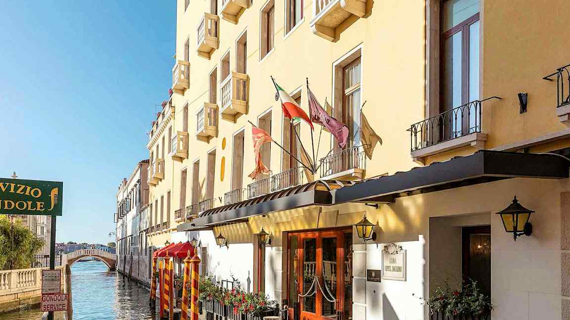 En gul bygning med balkonger langs en kanal, med en gangbro i bakgrunnen. (AI-generert tekst)