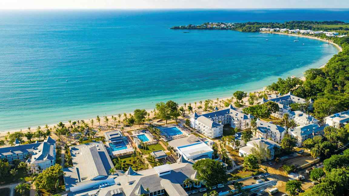 Bilde av en kyststrekning med turkis vann, en hvit sandstrand og hotellbygninger i nærheten. (AI-generert tekst)