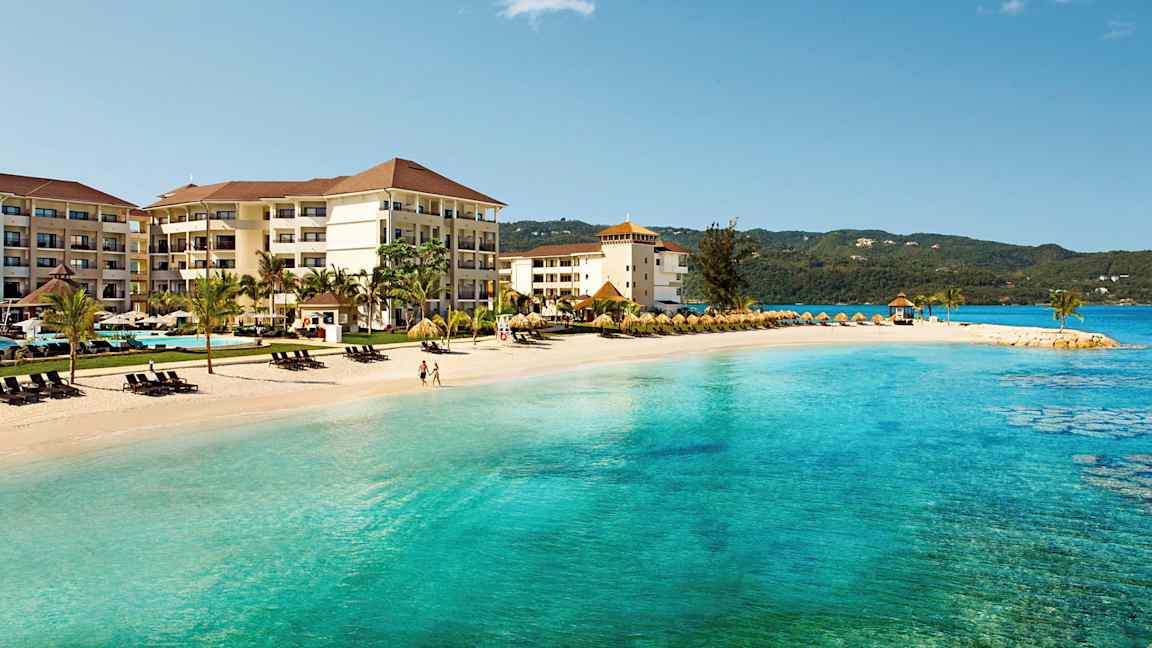 På stranden ligger et hotell med flere etasjer og palmetrær, ved siden av turkist hav og solsenger. (AI-generert tekst)