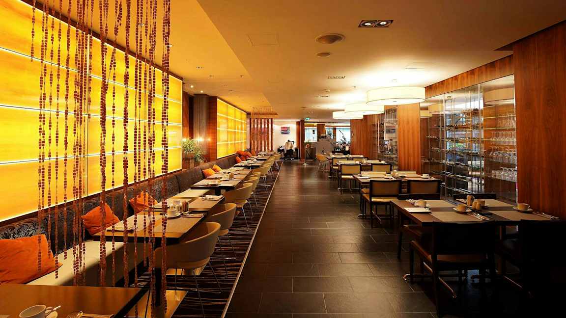 Bilde av en restaurant med moderne møbler, varme farger og rekke med dekkede bord. En hylle med glass er synlig til høyre. (AI-generert tekst)