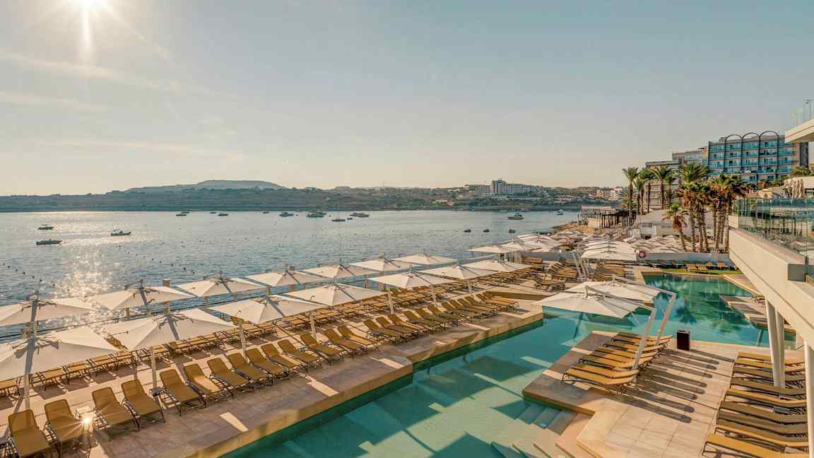 Utsikt over et solfylt bassengområde med solstoler og parasoller, med havet og båter i bakgrunnen. (AI-generert tekst)