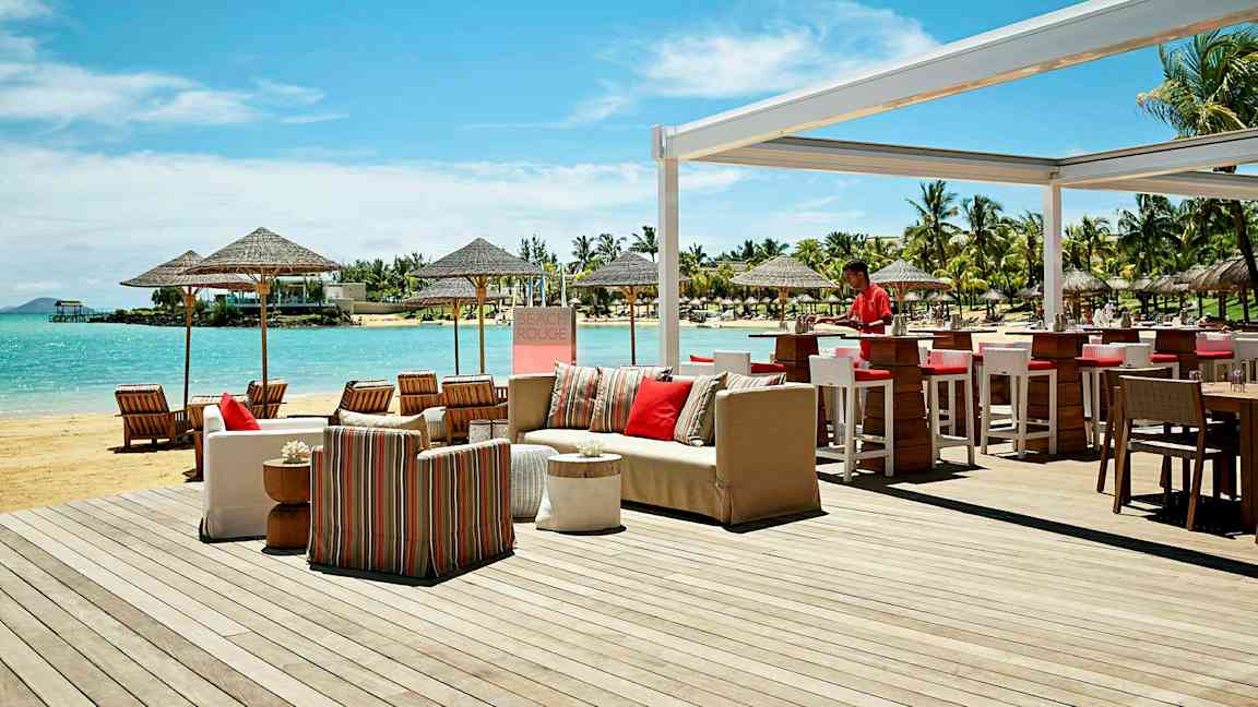Utendørs serveringsområde ved stranden med bar og sittegrupper, samt utsikt over havet og omkringliggende palmer. (AI-generert tekst)