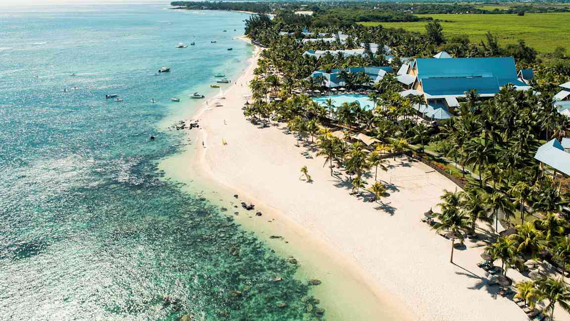 Utsikt over en kyst med en bred sandstrand, turkis hav og et bassengområde omgitt av palmer og hotellbygninger. (AI-generert tekst)