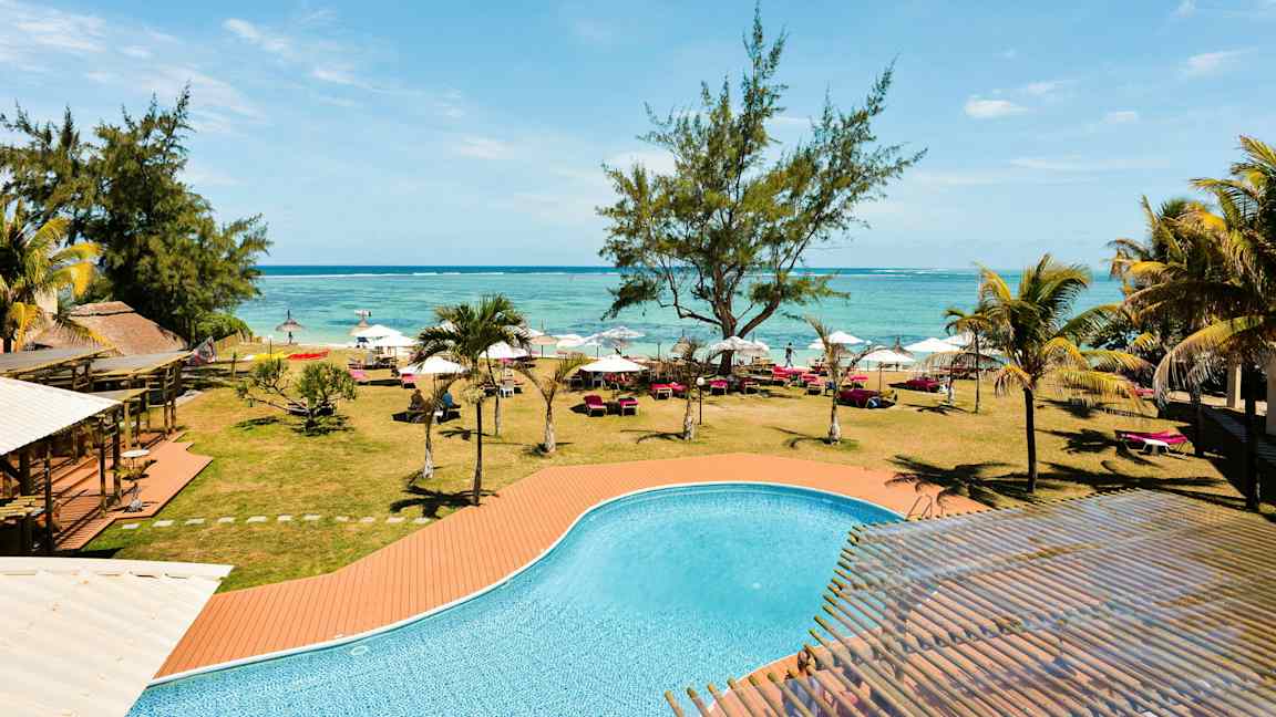 Et basseng med turkist vann foran en plen med trær, solsenger og parasoller langs en strand. (AI-generert tekst)