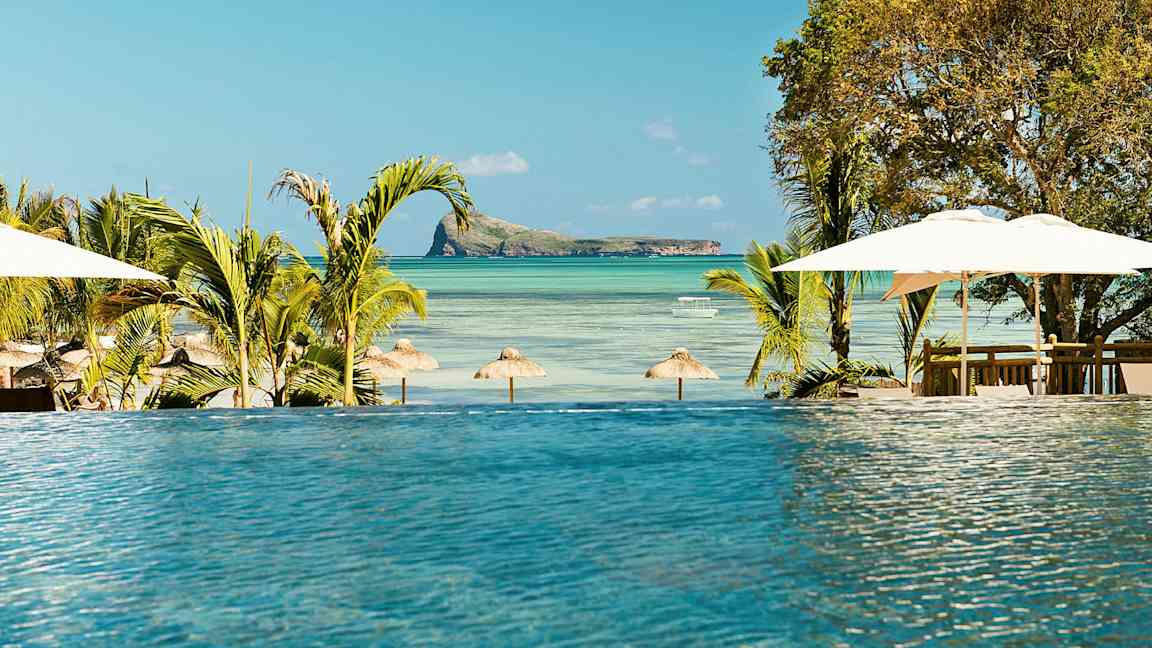 Et infinitybasseng med utsikt over en strand med parasoller og en liten øy i havet. (AI-generert tekst)