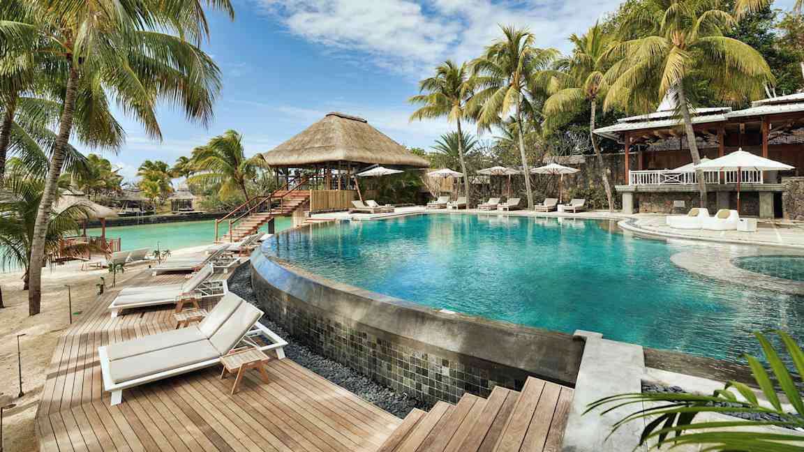 Et basseng omgitt av solsenger og parasoller, med palmer og en bygning med stråtak i bakgrunnen. (AI-generert tekst)