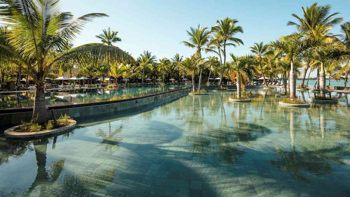 Et stort svømmebasseng omgitt av palmer under en klar blå himmel. (AI-generert tekst)