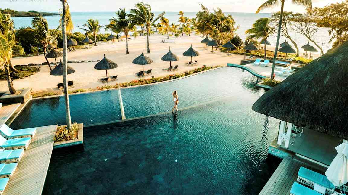 En tropisk strand med palmer og parasoller ved havet, et infinity-basseng i forgrunnen. (AI-generert tekst)