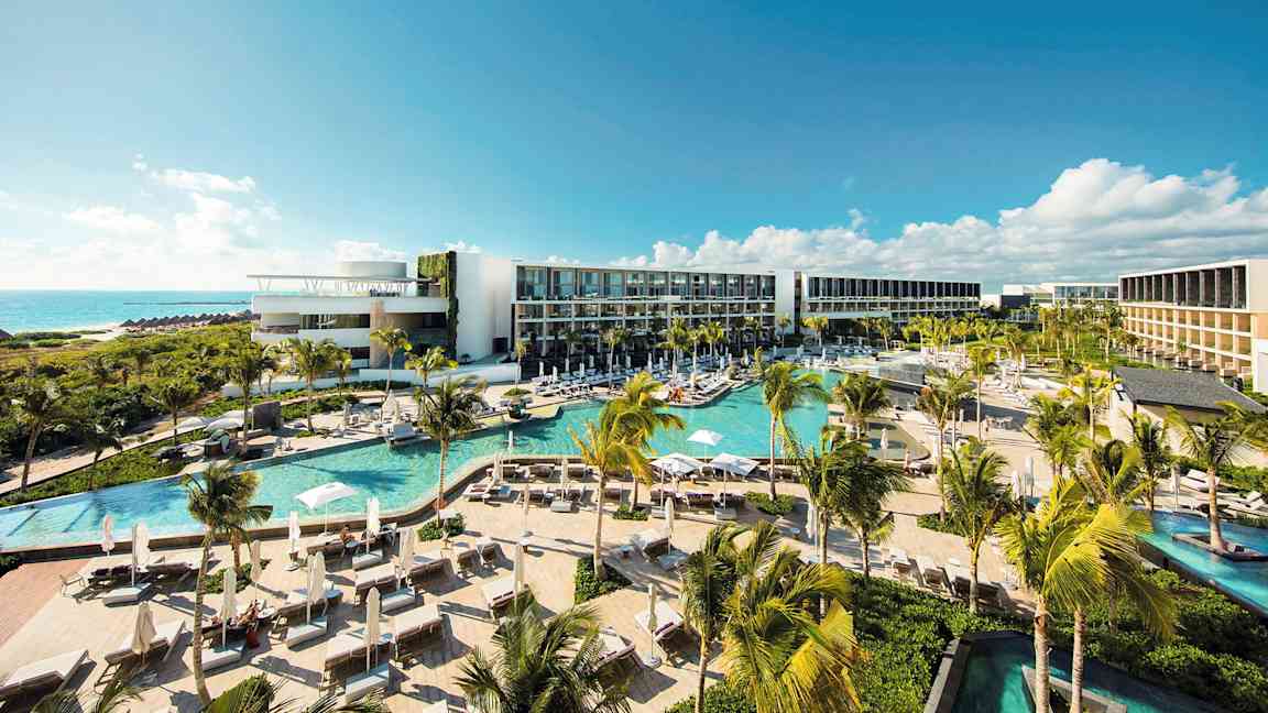 Et moderne hotellkompleks med flere bassenger, frodig vegetasjon og palmer, beliggende ved kysten med utsikt over havet under en klar blå himmel. (AI-generert tekst)