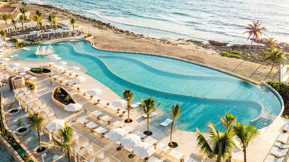 Et stort basseng omgitt av solsenger og parasoller ligger ved stranden med utsikt over havet. (AI-generert tekst)