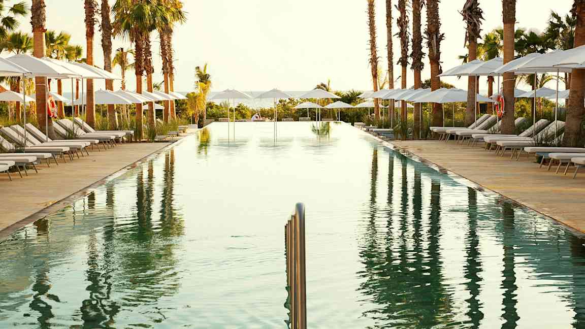 Et avlangt svømmebasseng omgitt av solsenger med parasoller og palmer. (AI-generert tekst)