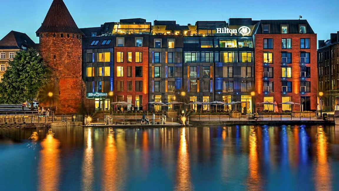En historisk murbygning ved en moderne hotell ved vannet om kvelden. (AI-generert tekst)