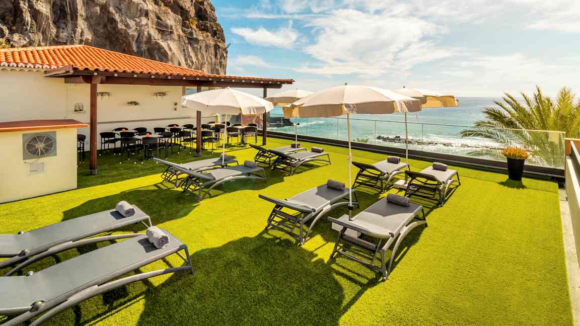 En terrasse med solsenger og parasoller på en gressplen, ved et lavt hus med tak i terrakottarødt. Utsikt over havet og en klippevegg i bakgrunnen. (AI-generert tekst)