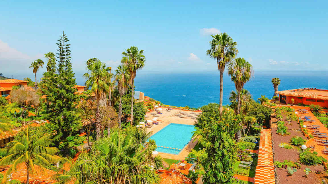 Utsikt over et bassengområde omgitt av frodighet og palmer, med havet i bakgrunnen. (AI-generert tekst)